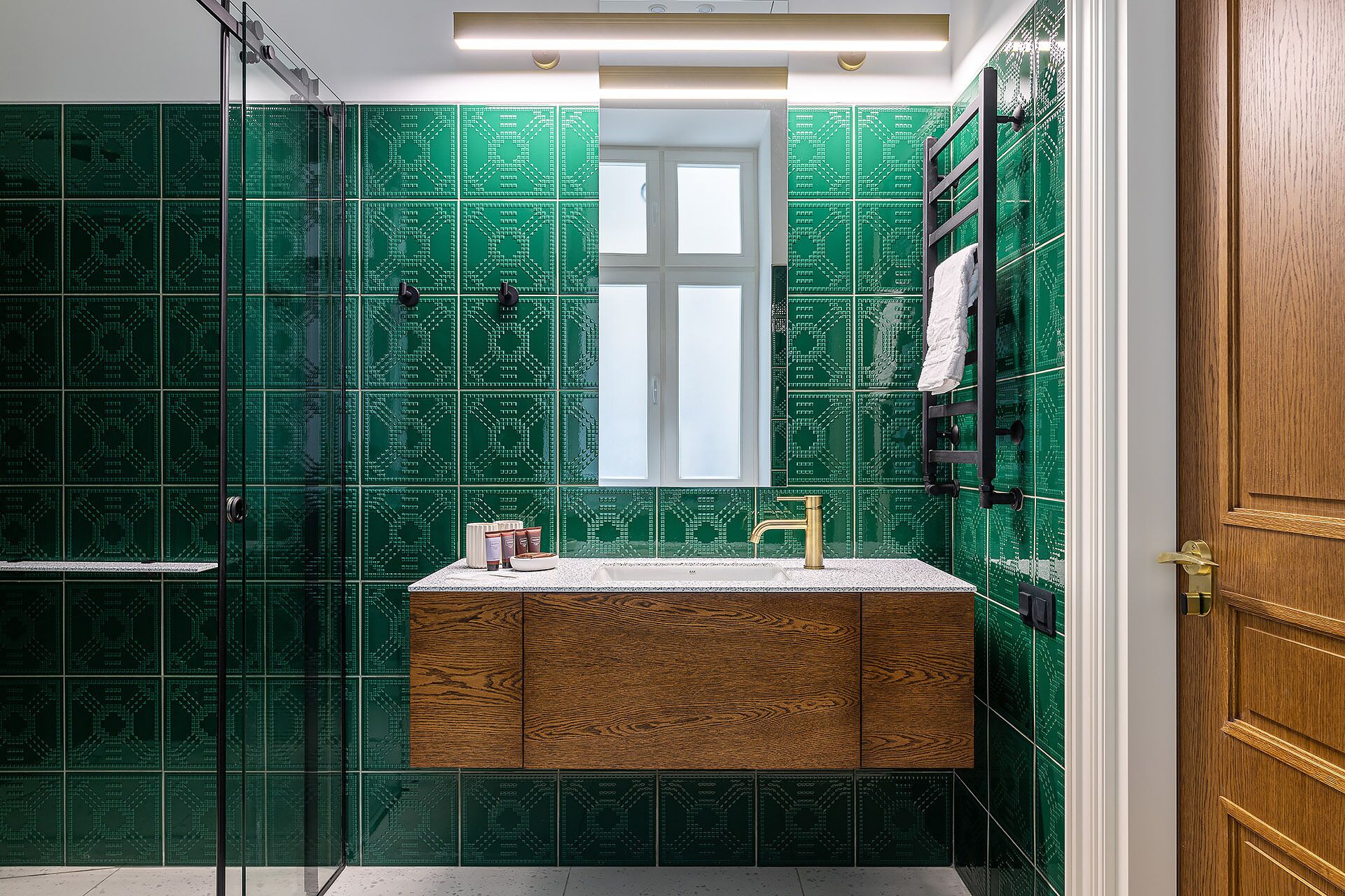 A bathroom with green patterned tiles, a wooden vanity, a gold faucet, and a window.