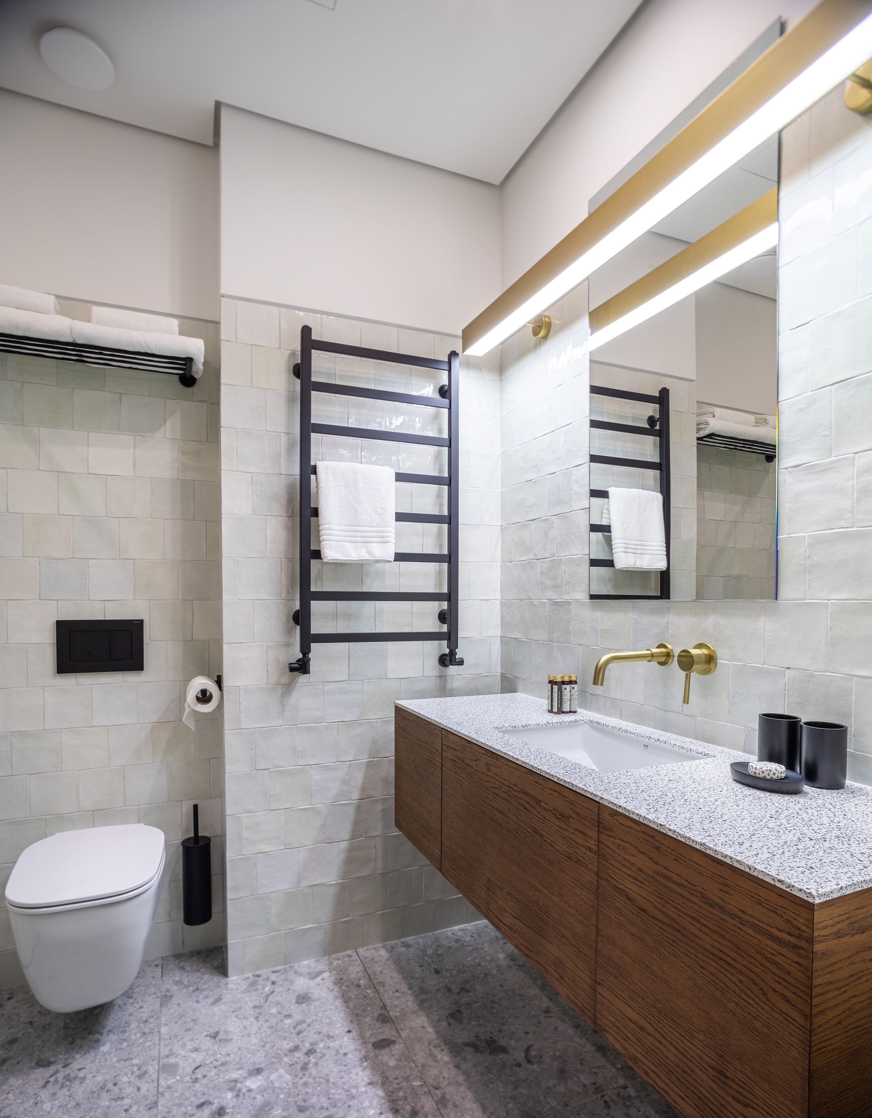 A modern bathroom with a white toilet, a sink with a wooden vanity, a towel rack, and a mirror with a light above it.
