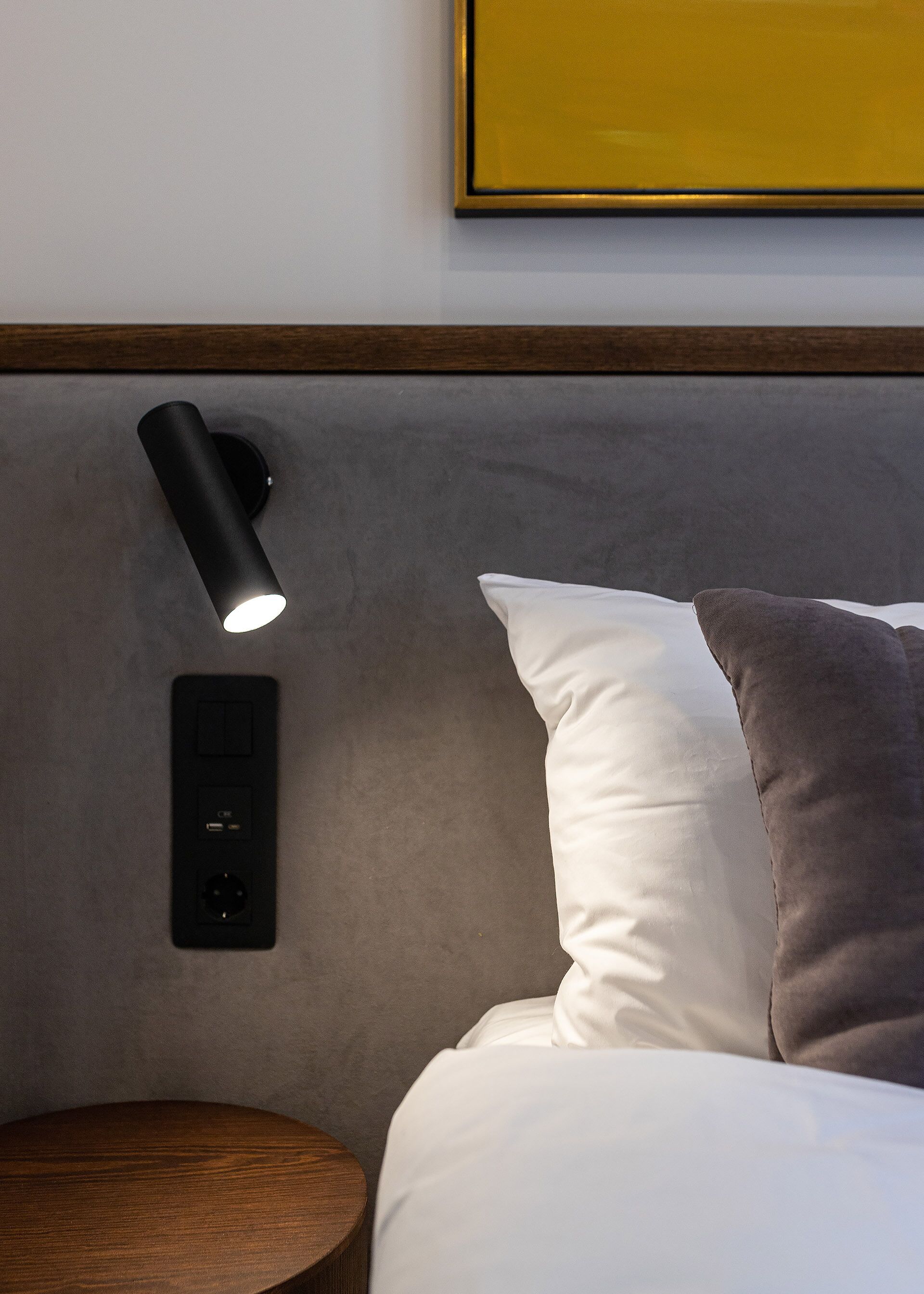 A modern bedroom with a headboard, a bedside lamp, a power outlet, and a wooden nightstand.