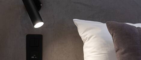 A modern bedroom with a headboard, a bedside lamp, a power outlet, and a wooden nightstand.