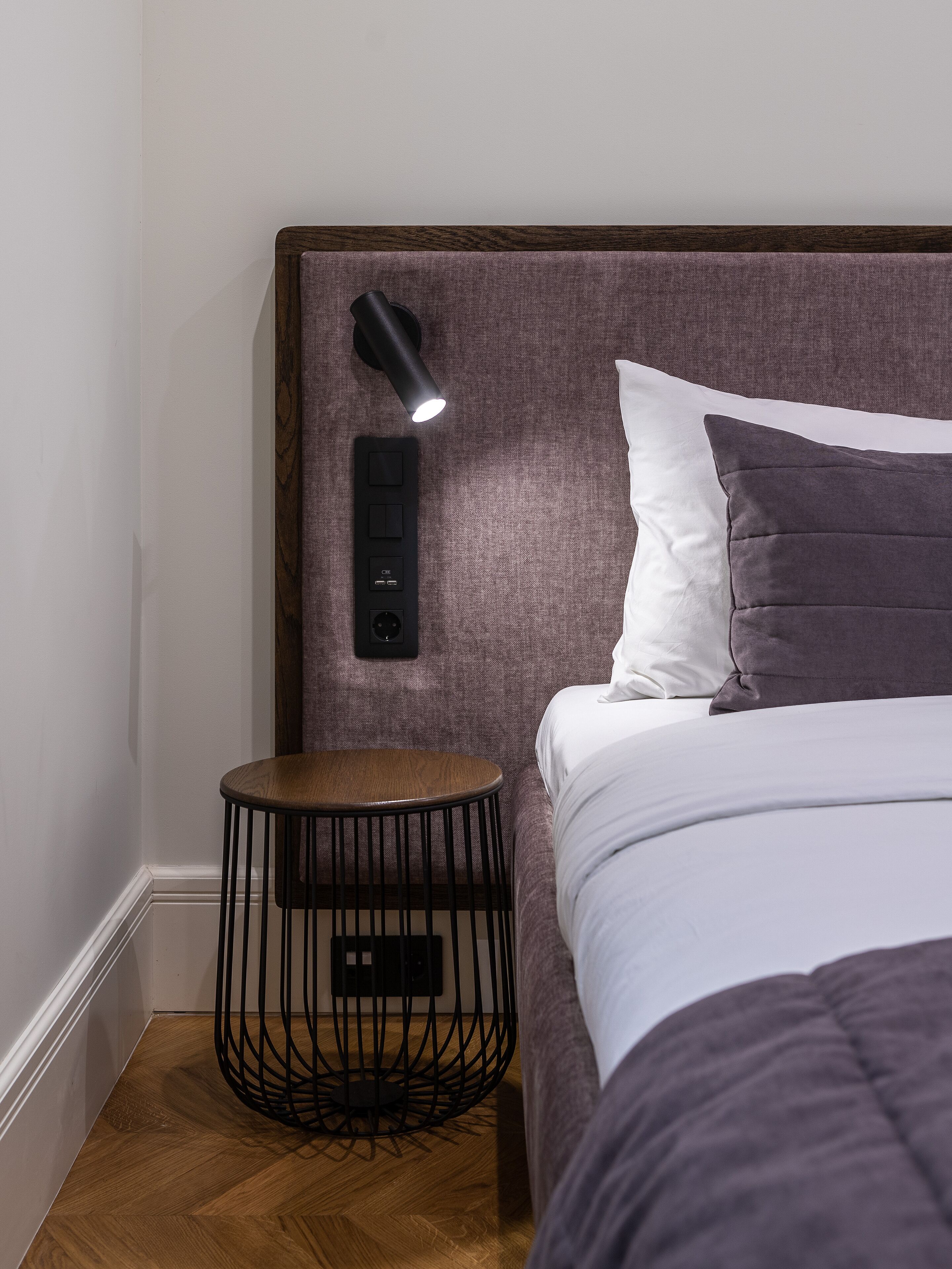 Una cama bien hecha con cabecera, una mesita redonda con base de metal negro y una lámpara de pared.