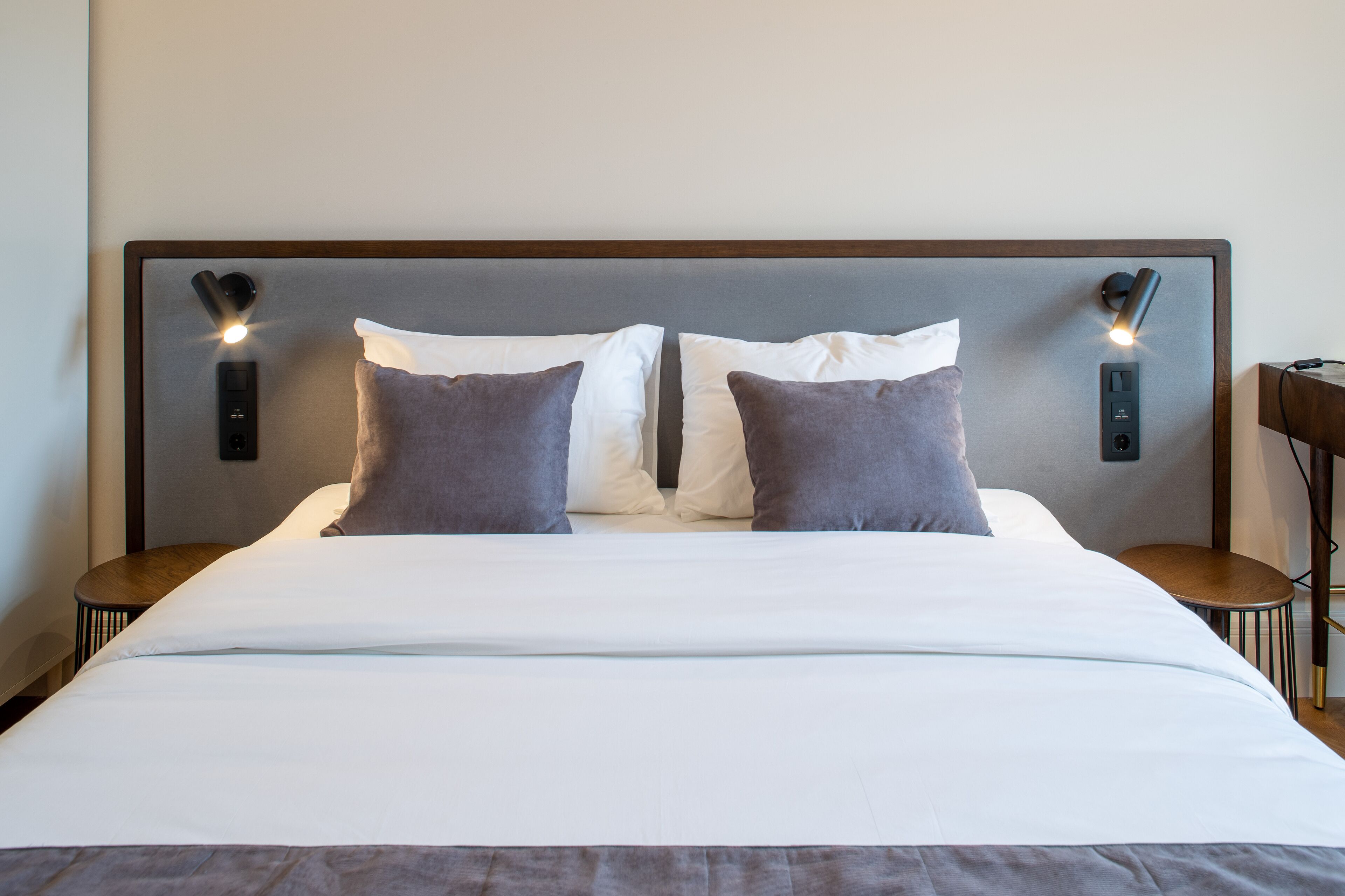 Una cama bien hecha con sábanas blancas y dos almohadas, un cabecero con iluminación integrada y una mesita de noche con lámpara.