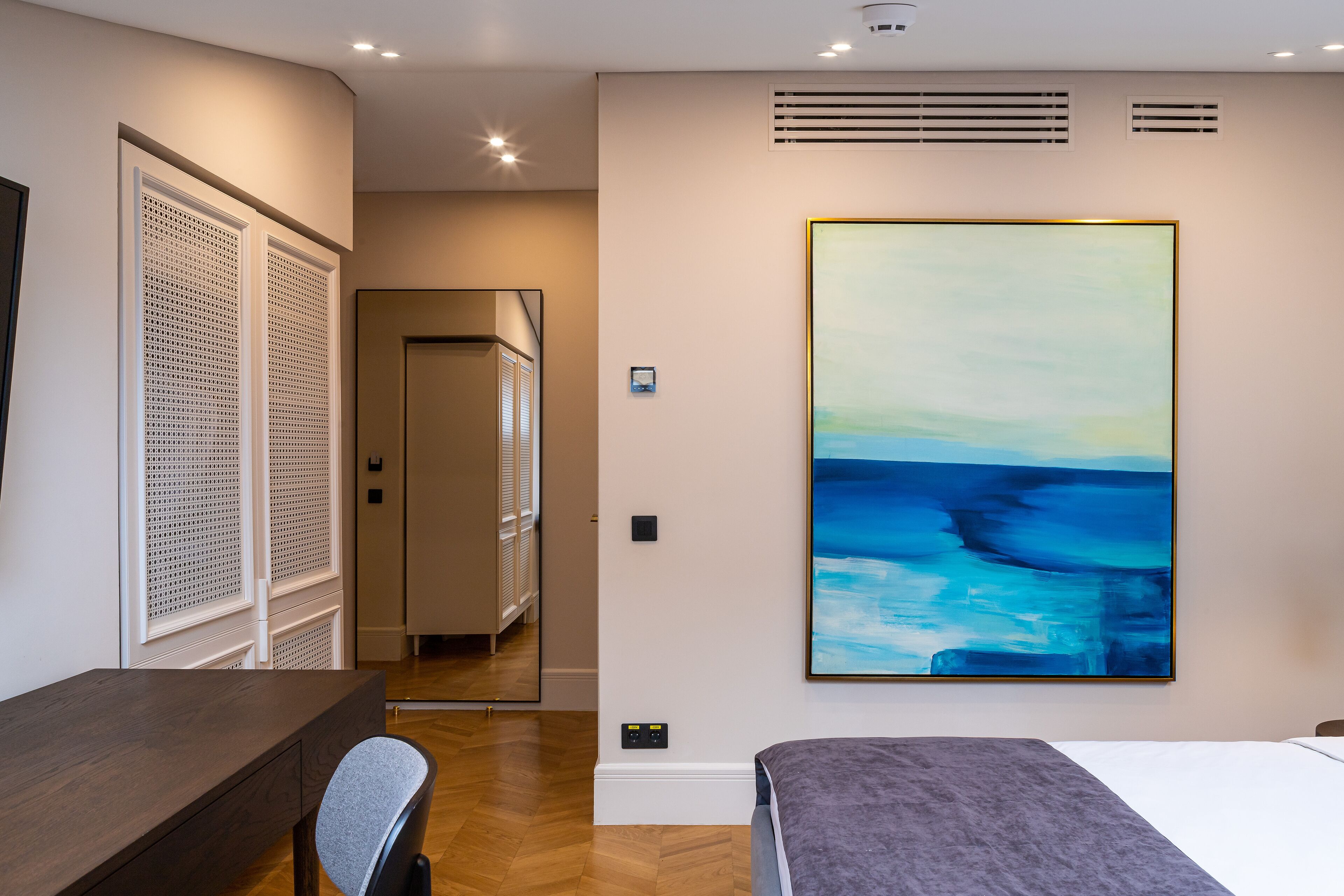 A modern hotel room with a wooden desk, a gray chair, a large bed, and a large abstract painting on the wall.
