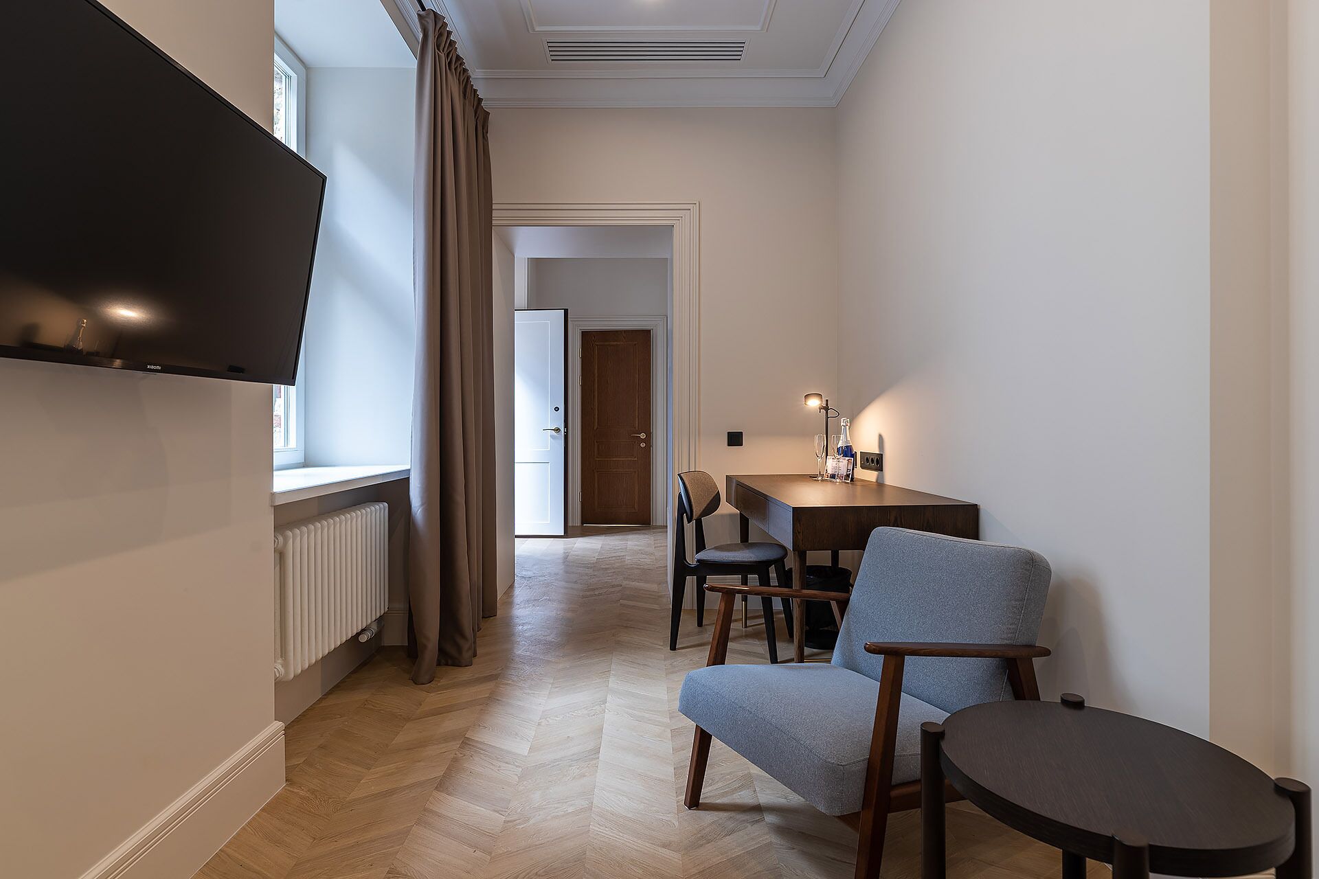 A modern hotel room with a flat-screen TV, a dining area with a table and chairs, and a large window with curtains.
