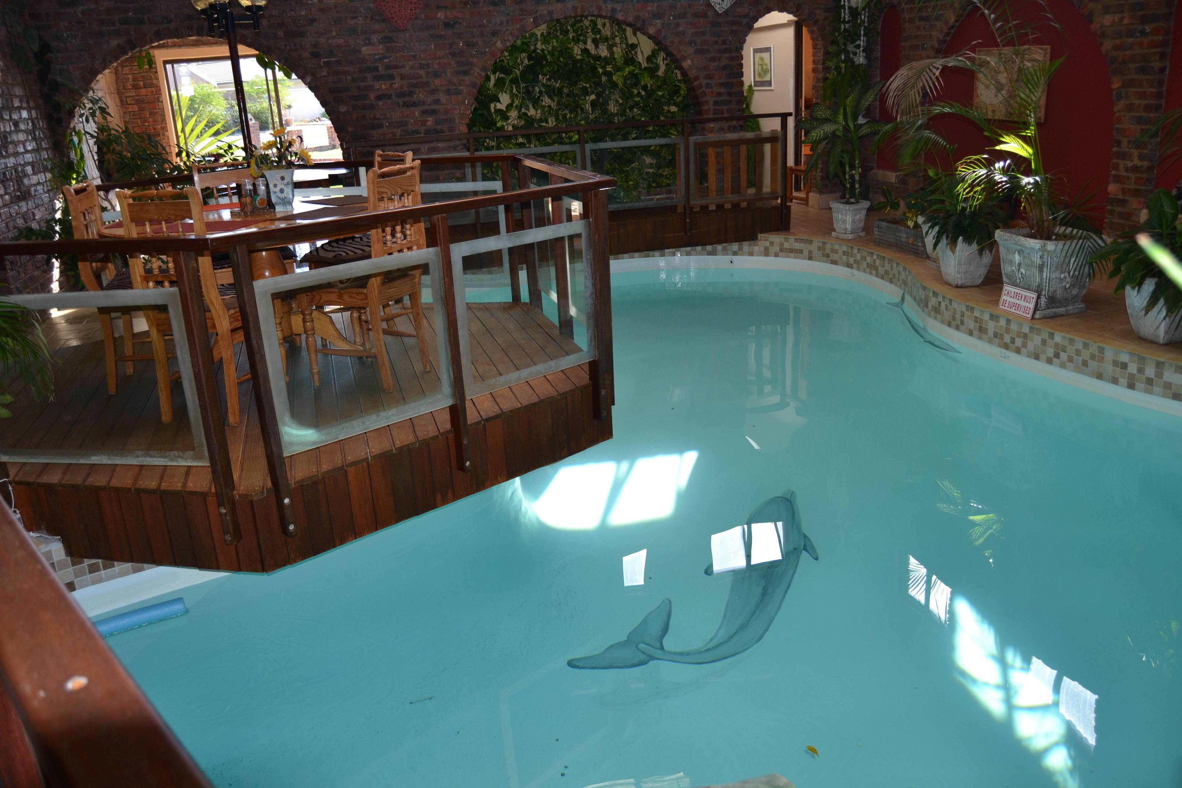Una piscina interior con un mural de delfines, una terraza de madera y plantas en macetas.
