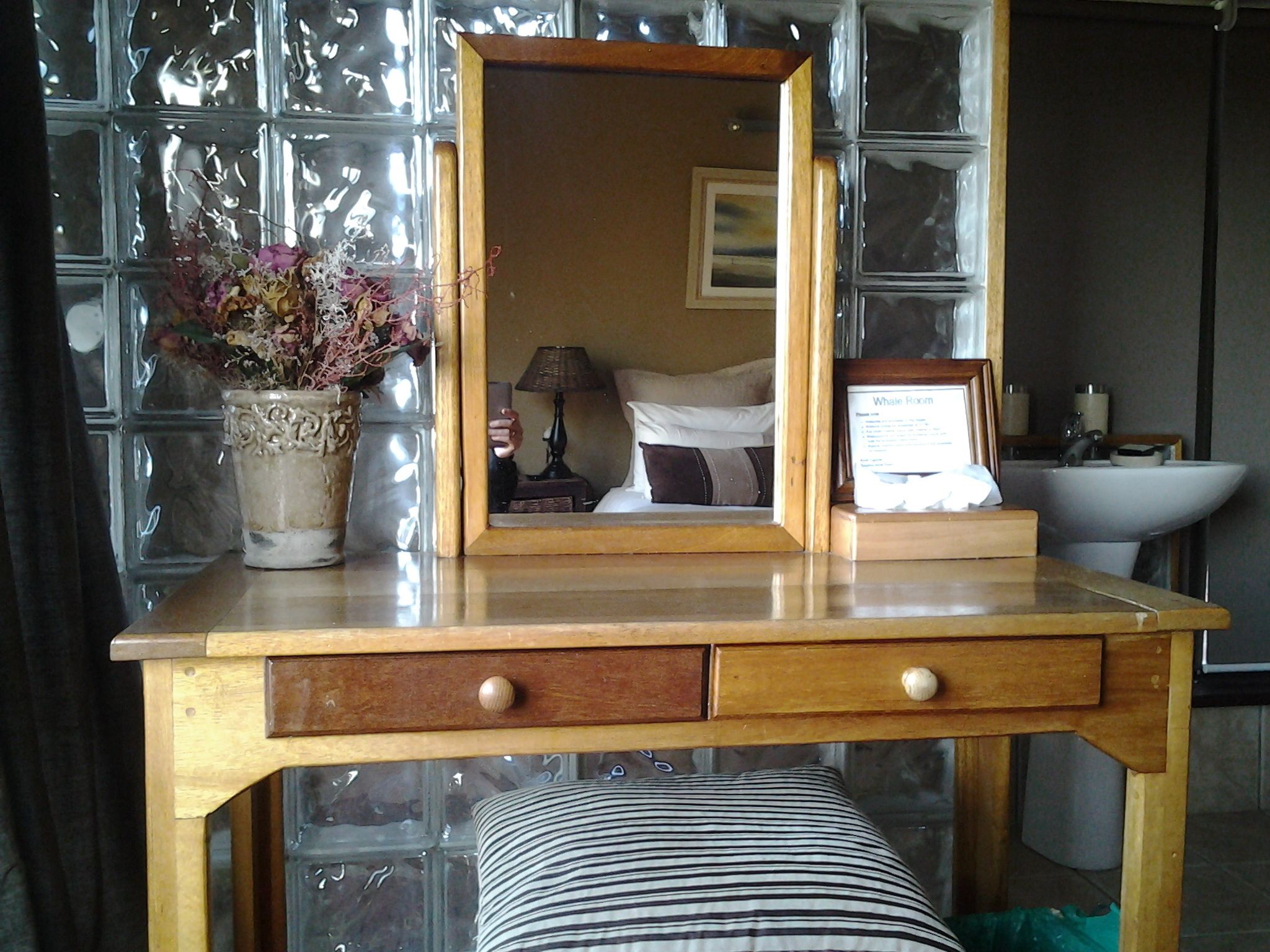 A wooden vanity with a mirror, a vase with flowers, and a bedside table with a lamp.