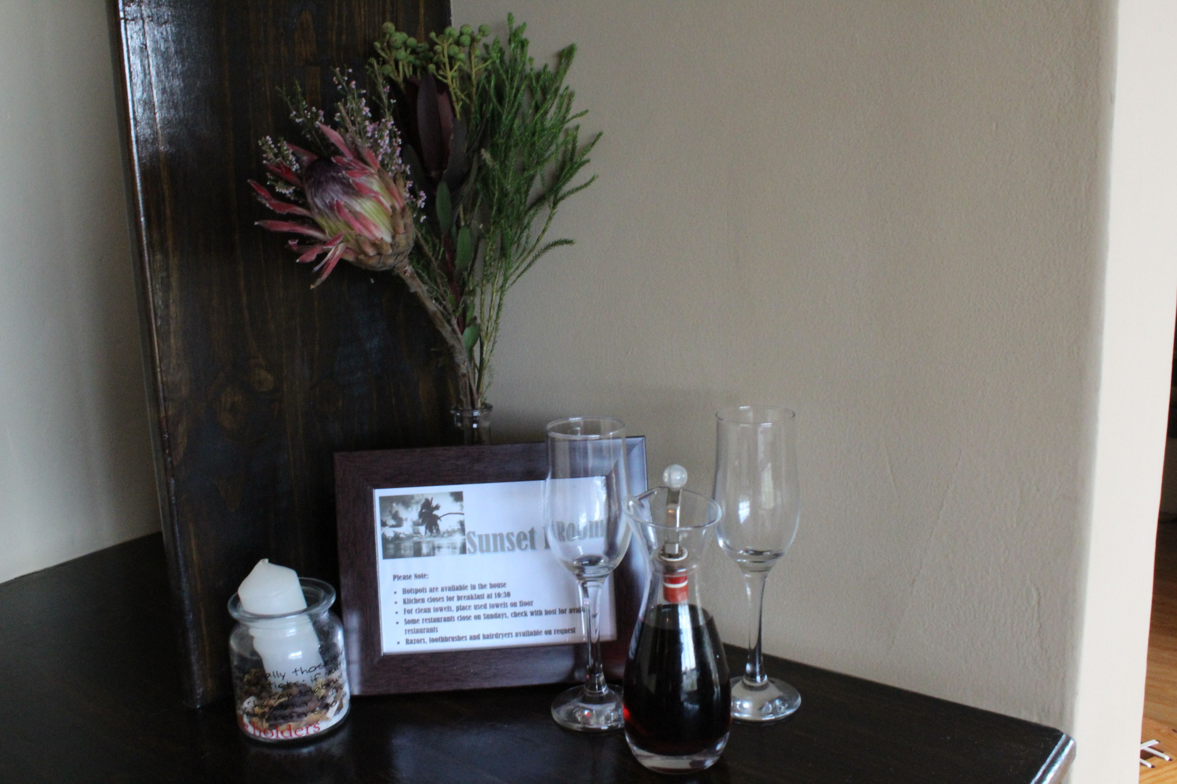 A table with a framed menu, two wine glasses, a bottle of wine, a small jar with a candle, and a vase with flowers.