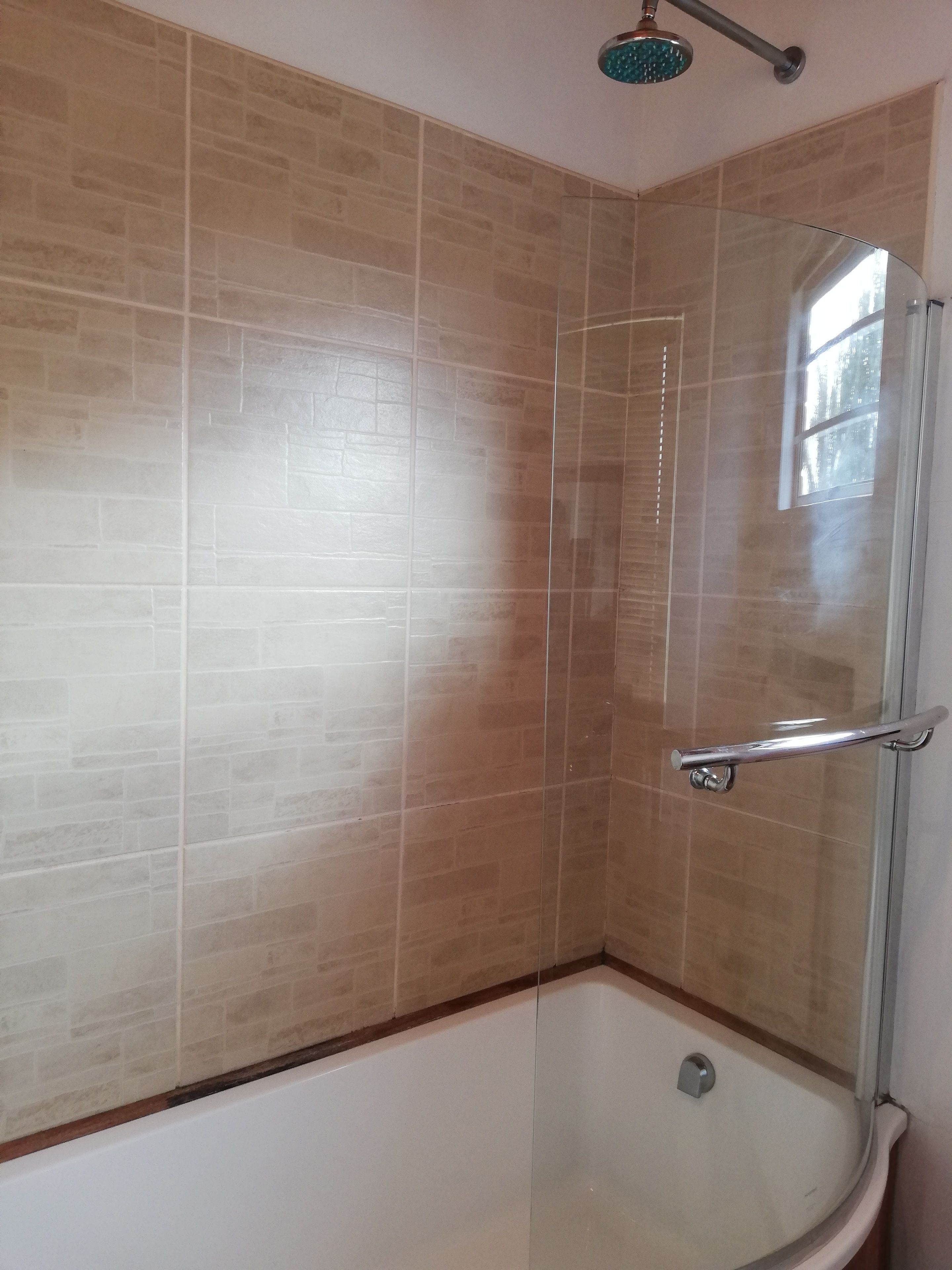 Una ducha con puerta de vidrio, paredes con azulejos color beige y una ventana.