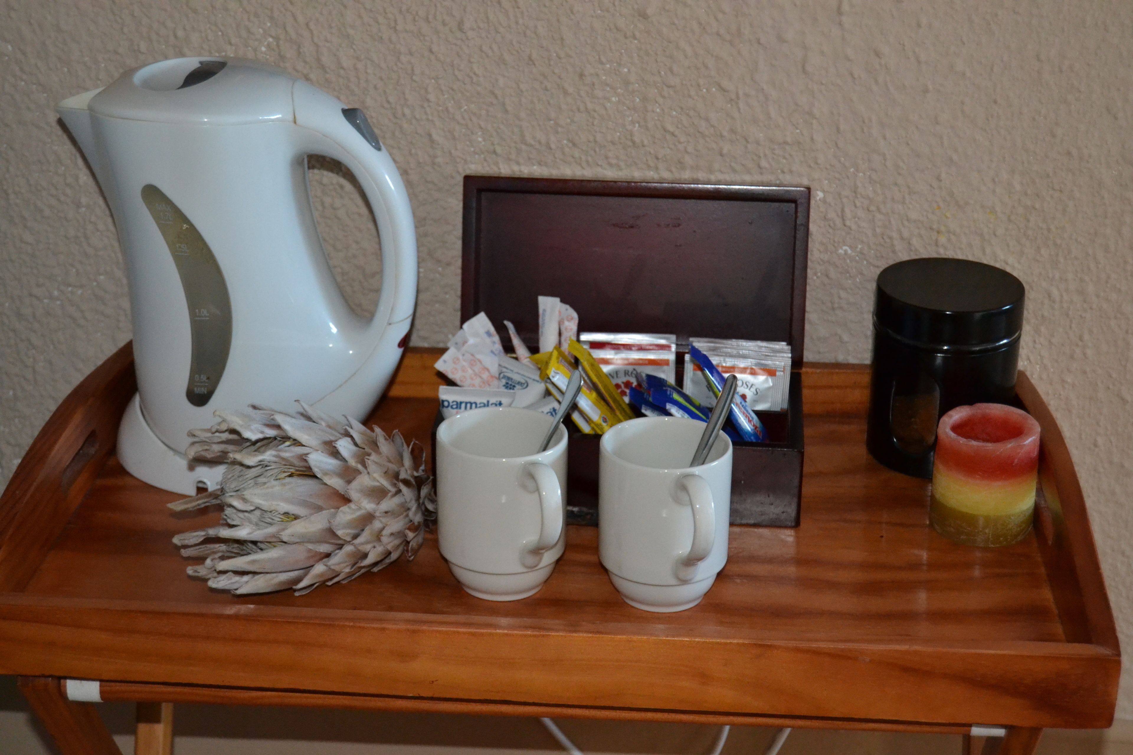 A wooden tray with a white electric kettle, two white mugs, a small black container, a colorful candle, and a box of tissues.