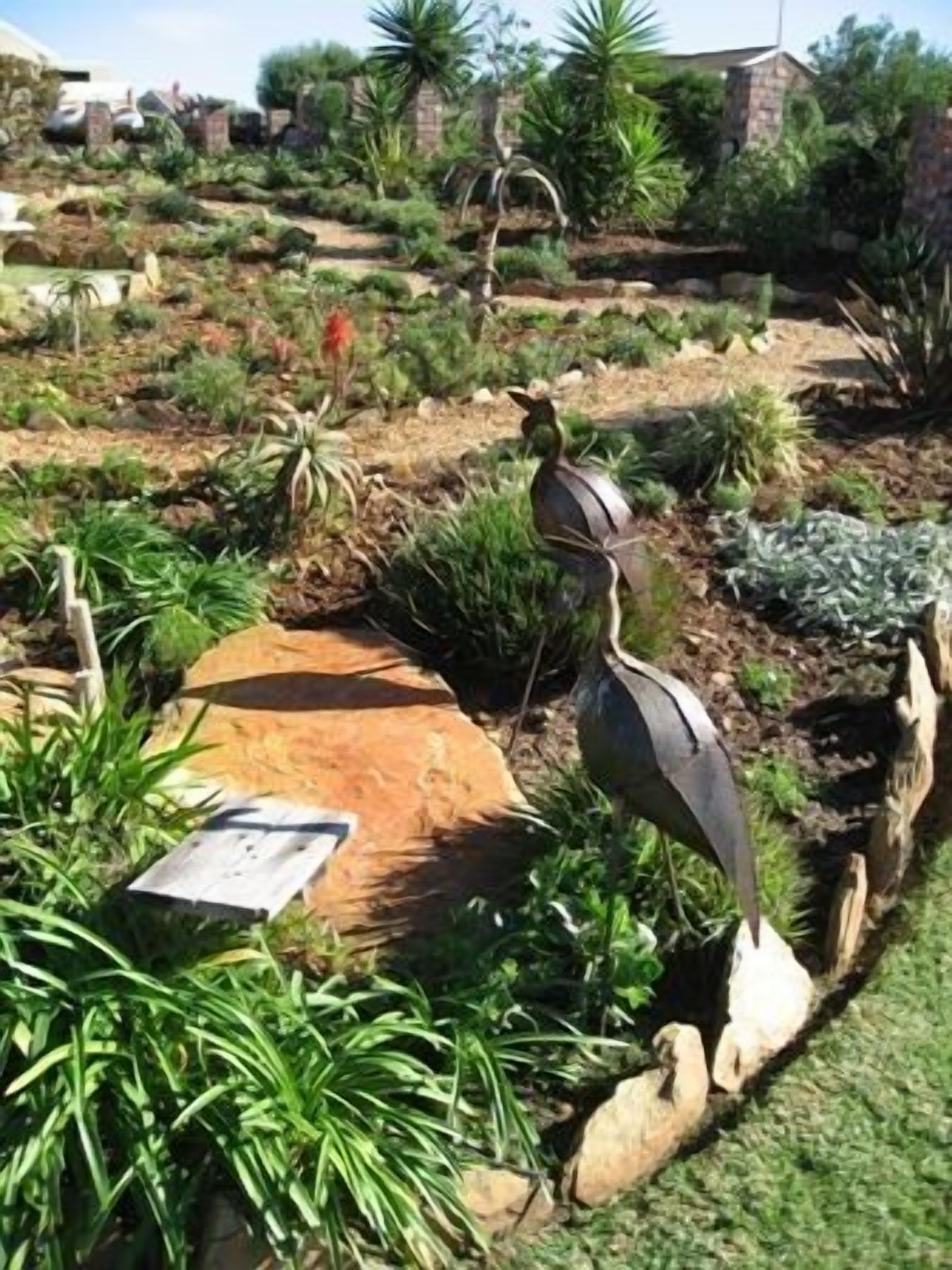 Un jardín con una escultura de pájaro, diversas plantas y un sendero de piedra.