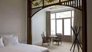 A hotel room with a bed, a small table with chairs, a window with curtains, and a wooden archway. - Wu Wei (Kortrijk)