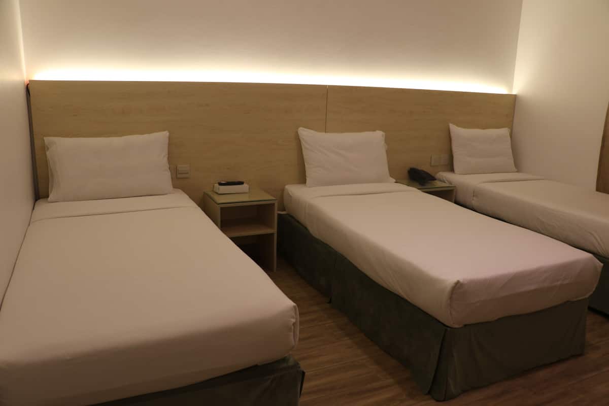 A hotel room with three single beds, a bedside table with a lamp, and a small shelf.