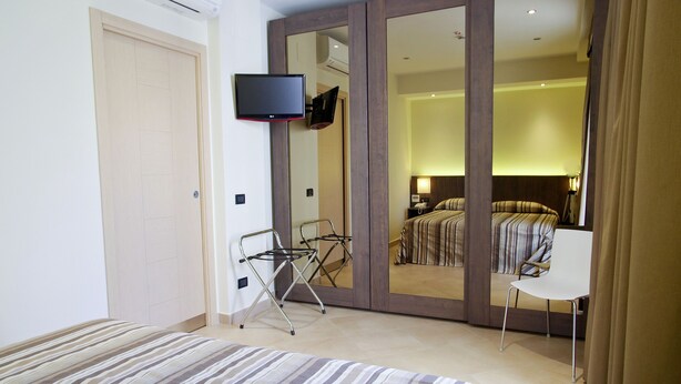 Habitación de hotel con cama, televisor montado en la pared, espejo, silla y mesita de noche con lámpara.