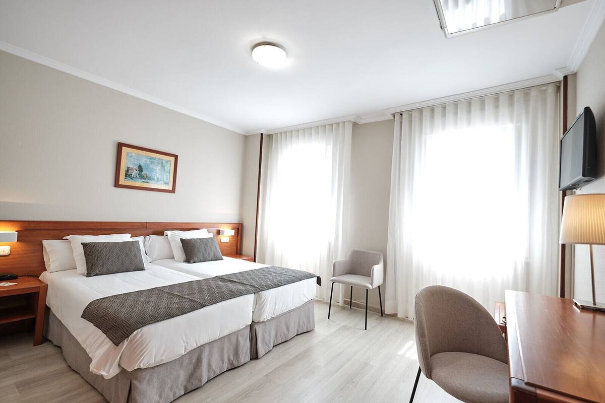 Una habitación de hotel con cama doble, un escritorio con silla, televisor y dos ventanas con cortinas.