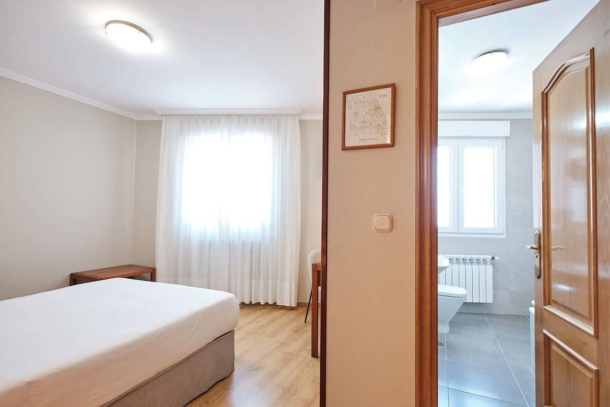Una habitación de hotel con cama, una mesita, una ventana con cortinas, una puerta y un baño visible a través de una puerta abierta.
