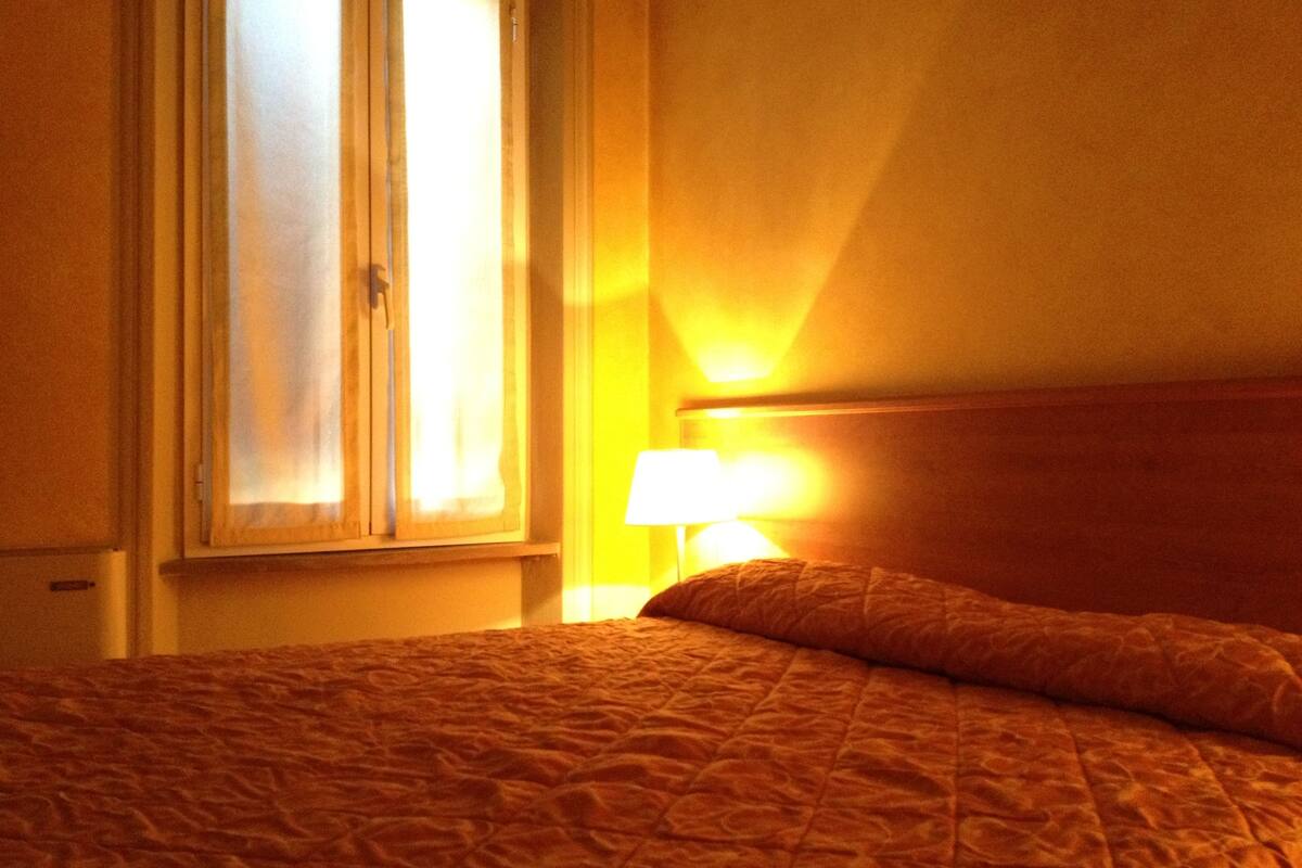 Una habitación de hotel con cama, ventana y mesita de noche con lámpara.