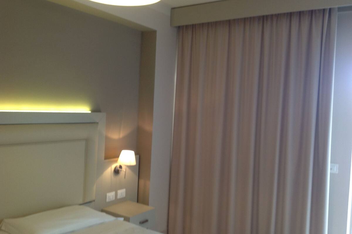 Habitació d'hotel amb un llit, una tauleta de nit, una làmpada i una finestra amb cortines.
