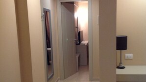 A hotel hallway with a tiled floor, a door leading to a bathroom, and a desk with a lamp.