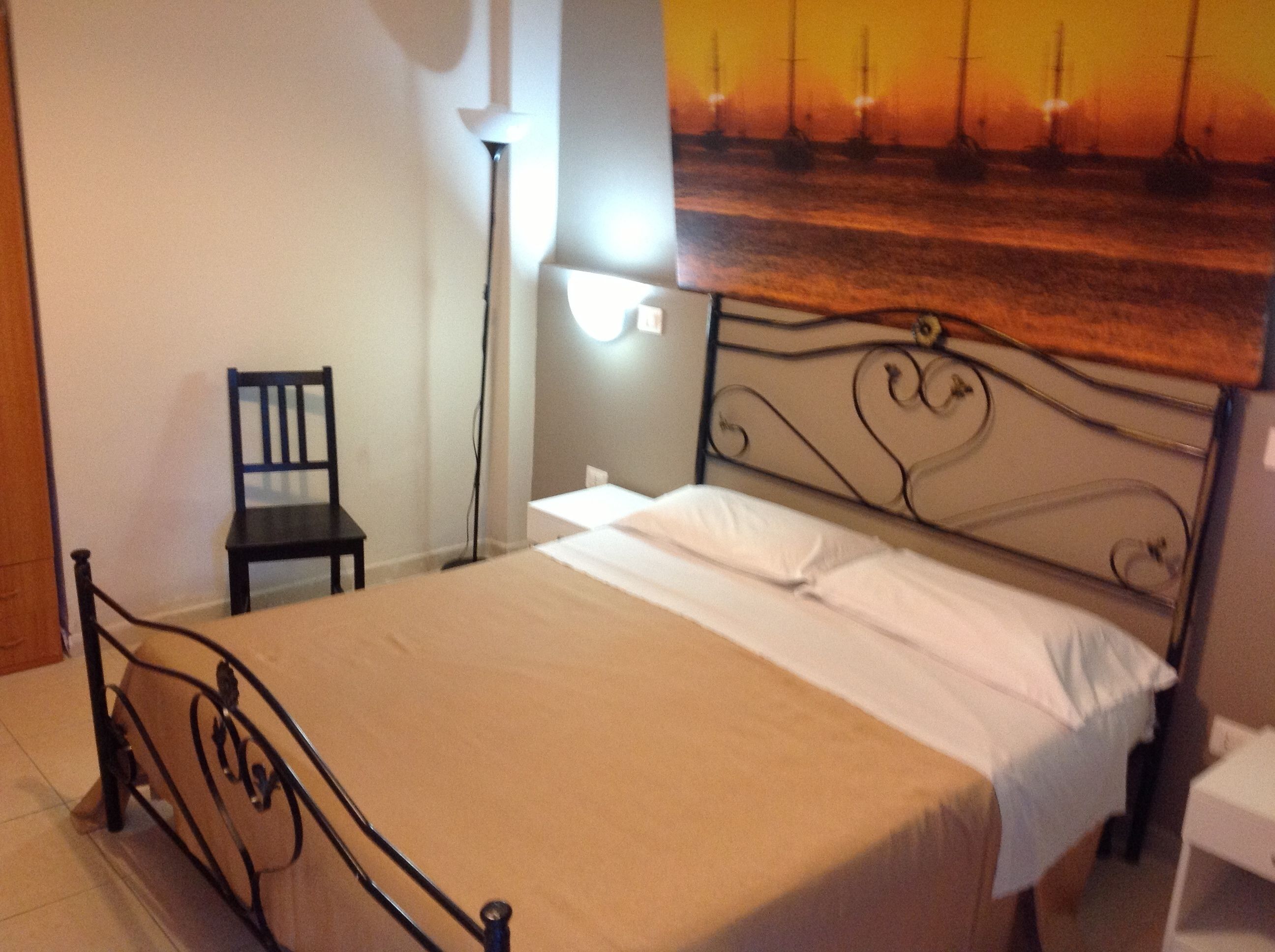 Una cama con estructura metálica, una silla, una lámpara de pared y un cuadro de un atardecer.