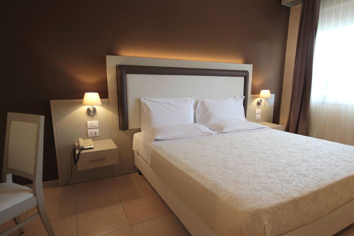 Habitació d'hotel amb un llit gran, dues taules de nit amb làmpades, una cadira i un telèfon.