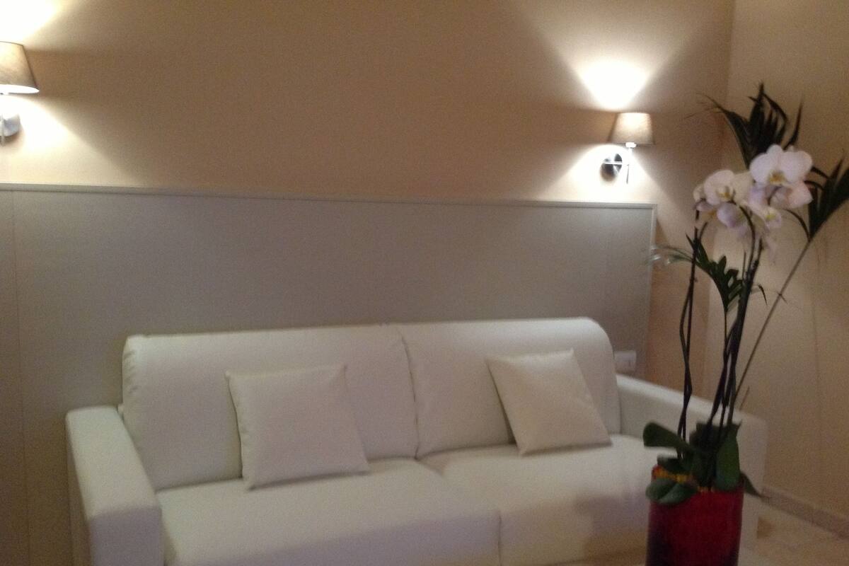 Un sofà blanc amb tres coixins, un vas vermell amb una planta i dues làmpades penjades a la paret.