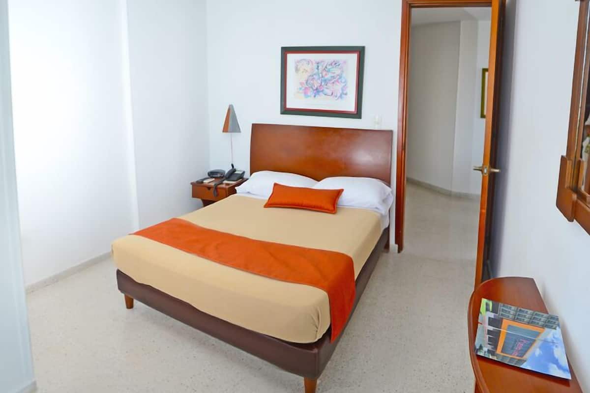 A hotel room with a large bed, a bedside table with a lamp, a framed picture on the wall, and a wooden door.
