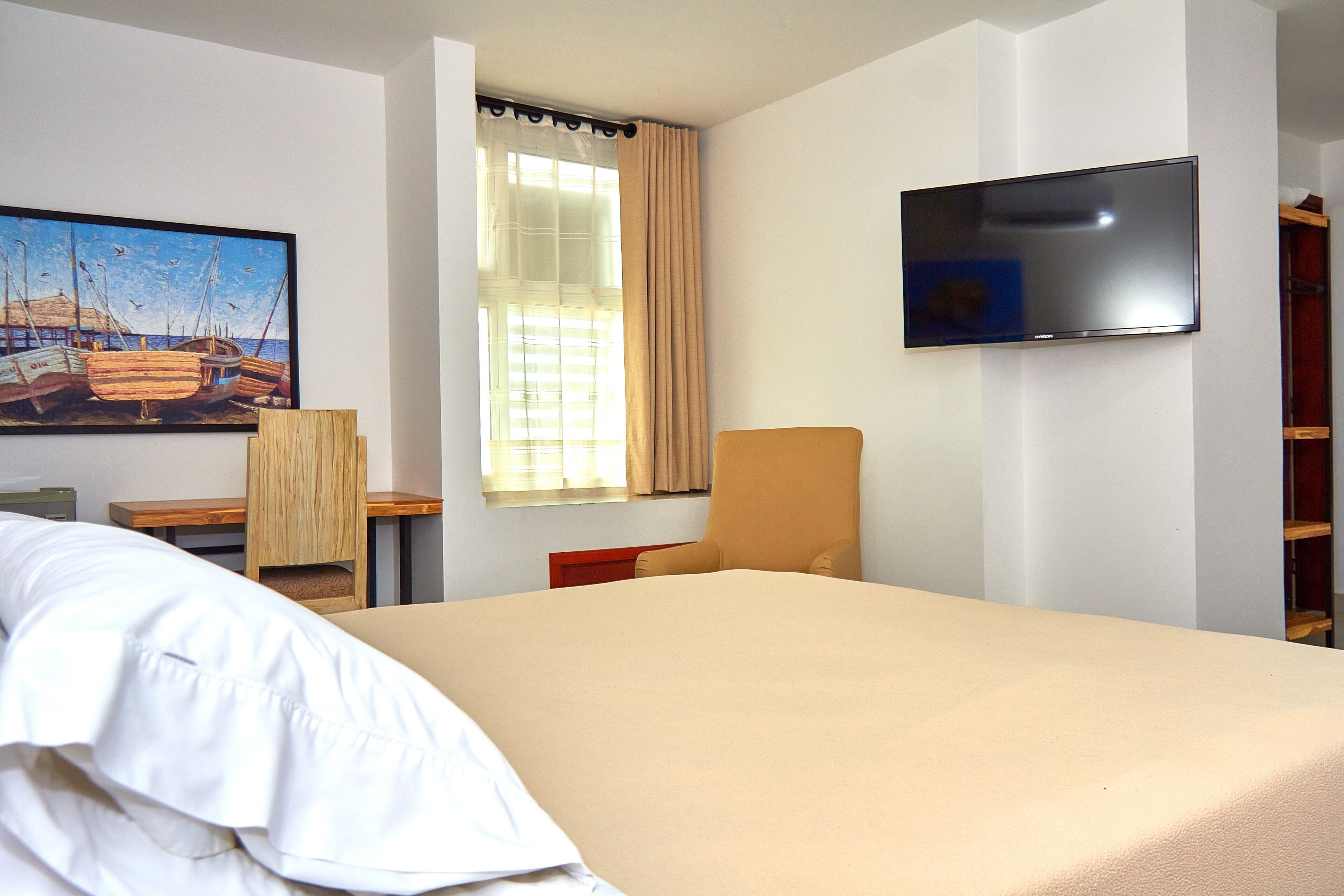 A hotel room with a bed, a TV mounted on the wall, a window with curtains, a painting on the wall, and a small table with a chair.