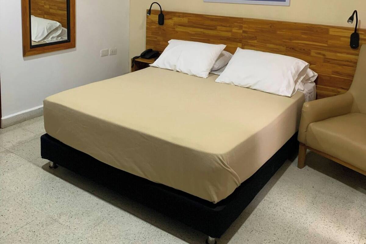 A hotel room with a bed, a chair, an air conditioning unit, a mirror, and a wall-mounted picture.