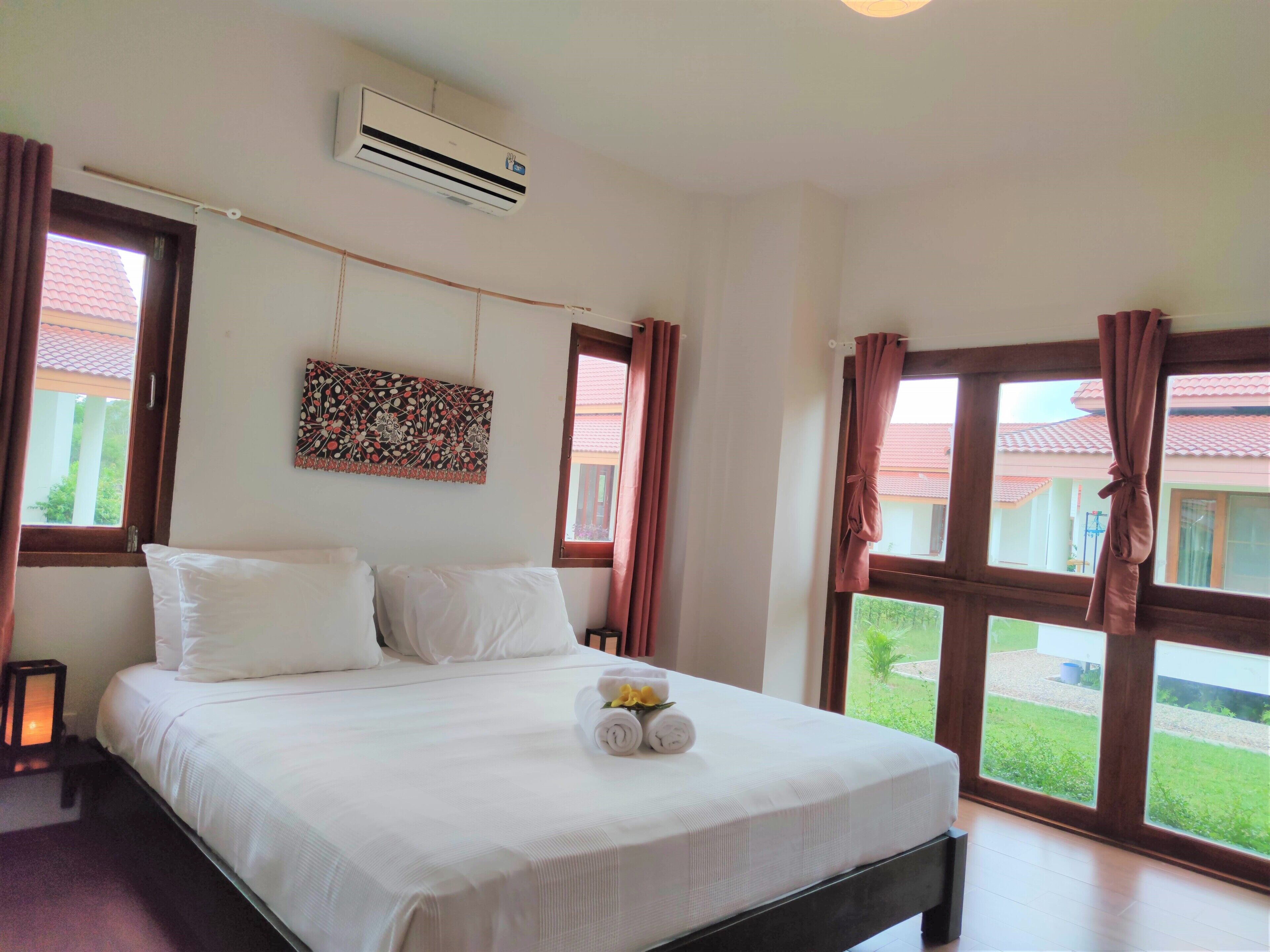 A hotel room with a large bed, an air conditioner, a window with curtains, and a view of a building with a red roof.