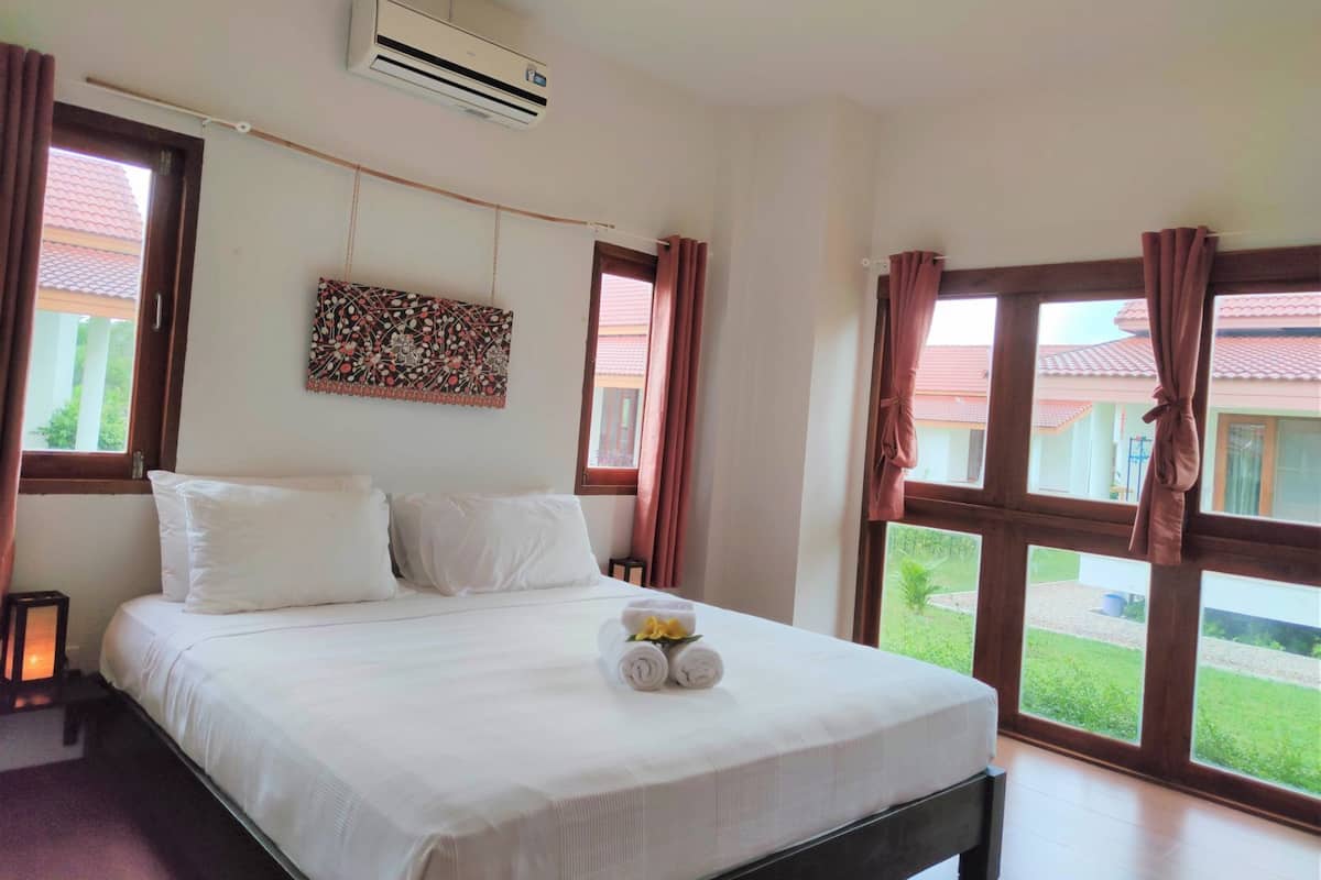 A hotel room with a bed, air conditioning unit, large windows, and a view of a building with a red roof.