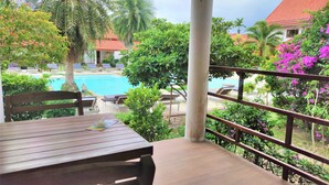 A balcony with a view of a swimming pool, palm trees, and a building with a red roof. - Armonia Village Resort and Spa (Chumphon)
