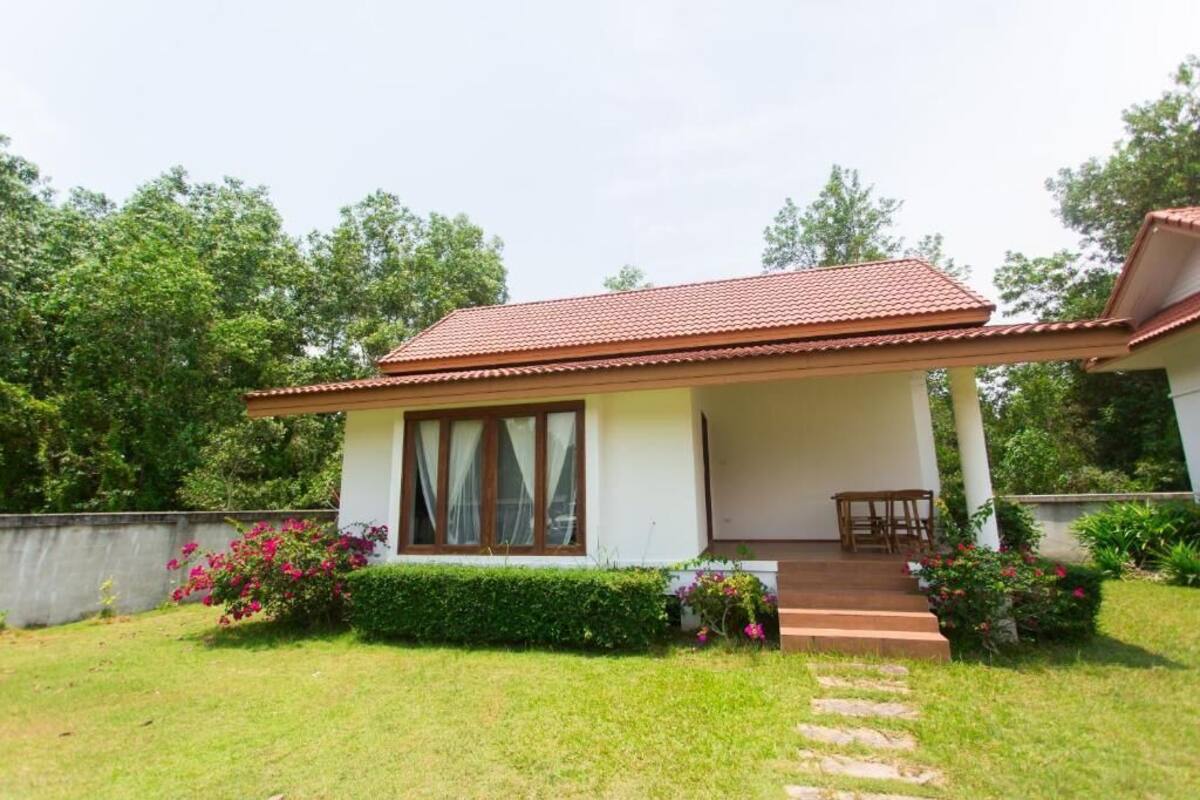 A single-story house with a tiled roof, a covered porch with a bench, and a small garden with flowering plants.