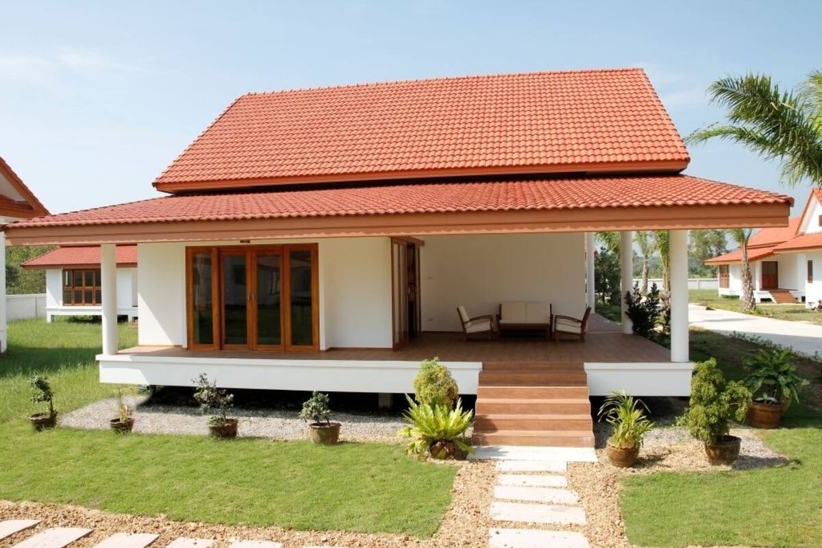 A two-story house with a red-tiled roof, a covered porch with seating, and a well-maintained lawn with potted plants.