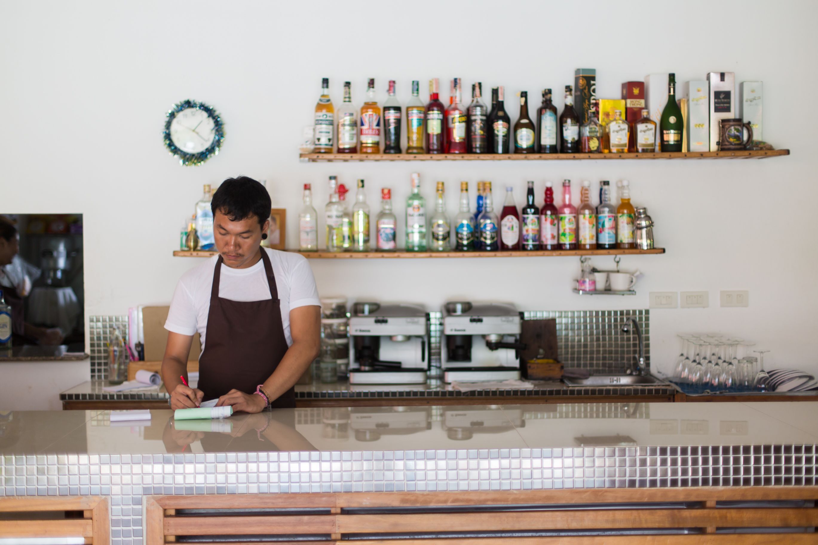 A bar with shelves of liquor bottles, a coffee machine, and a counter where a person is writing.