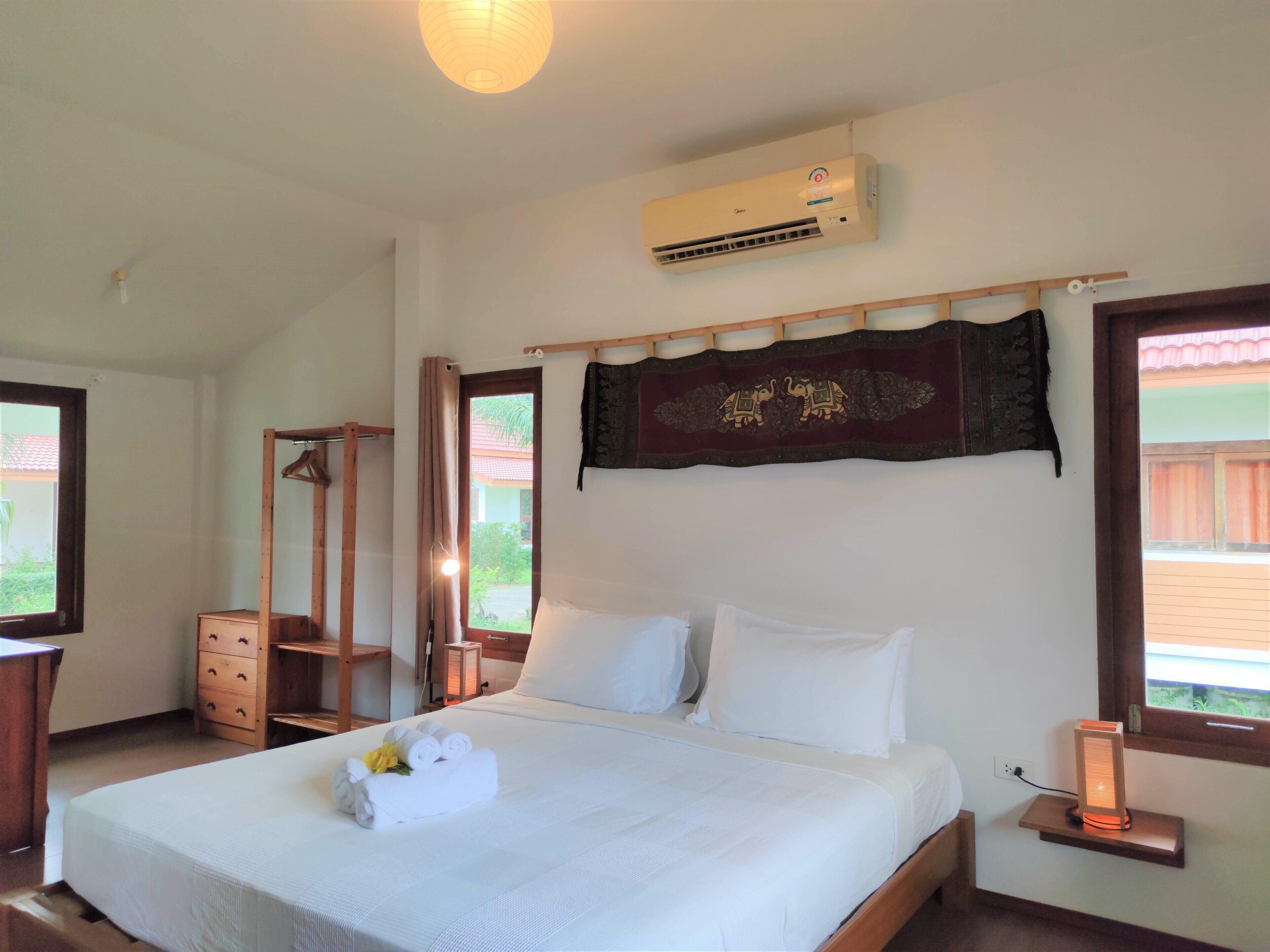 A hotel room with a bed, a wooden wardrobe, a mirror, an air conditioning unit, and a window with a view of greenery.