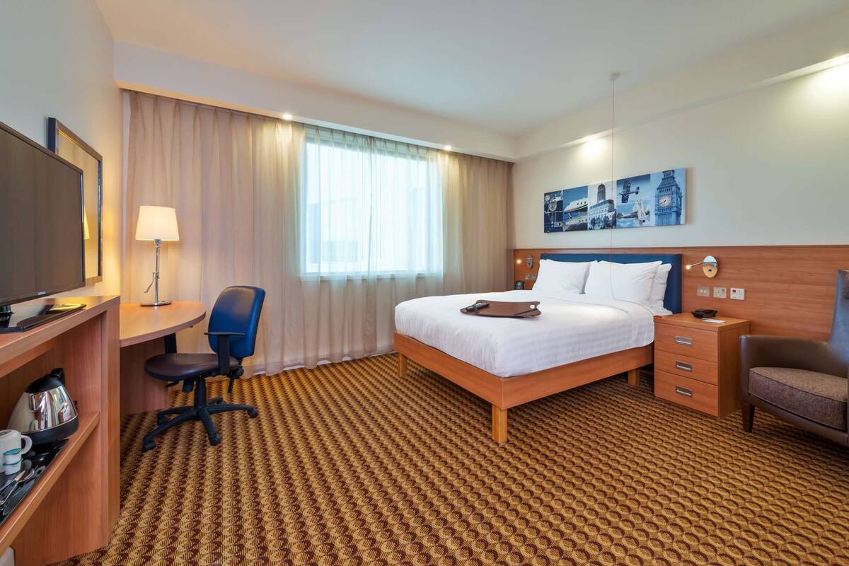 A hotel room with a bed, a desk with a chair, a television, a lamp, a nightstand, and a wall painting.