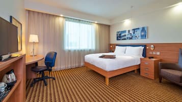 Habitación de hotel con cama, escritorio con silla, televisor, lámpara, mesita de noche y un cuadro en la pared.