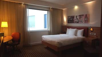 Una habitación de hotel con cama, escritorio con silla, lámpara, mesita de noche y un cuadro en la pared.