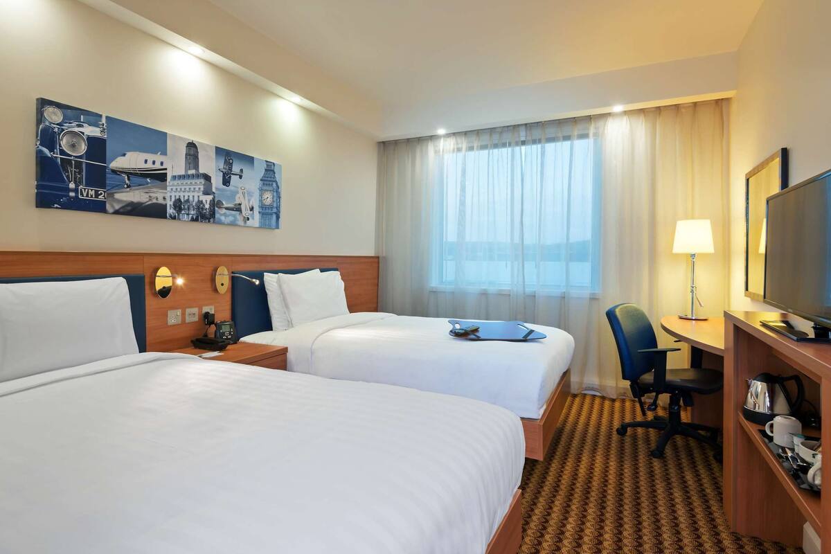 A hotel room with two beds, a desk, a chair, a TV, and a painting on the wall.