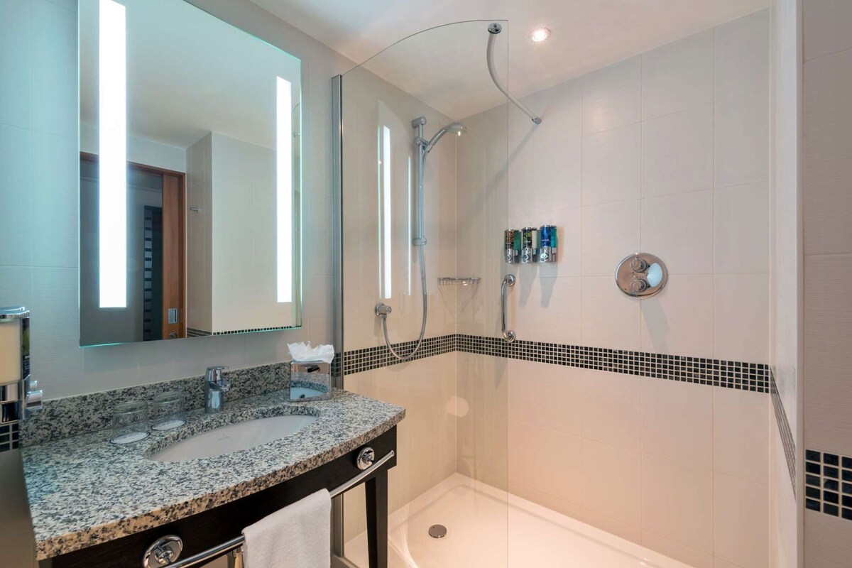 A bathroom with a granite countertop, a mirror, a shower, and a tiled wall with a black and white border.