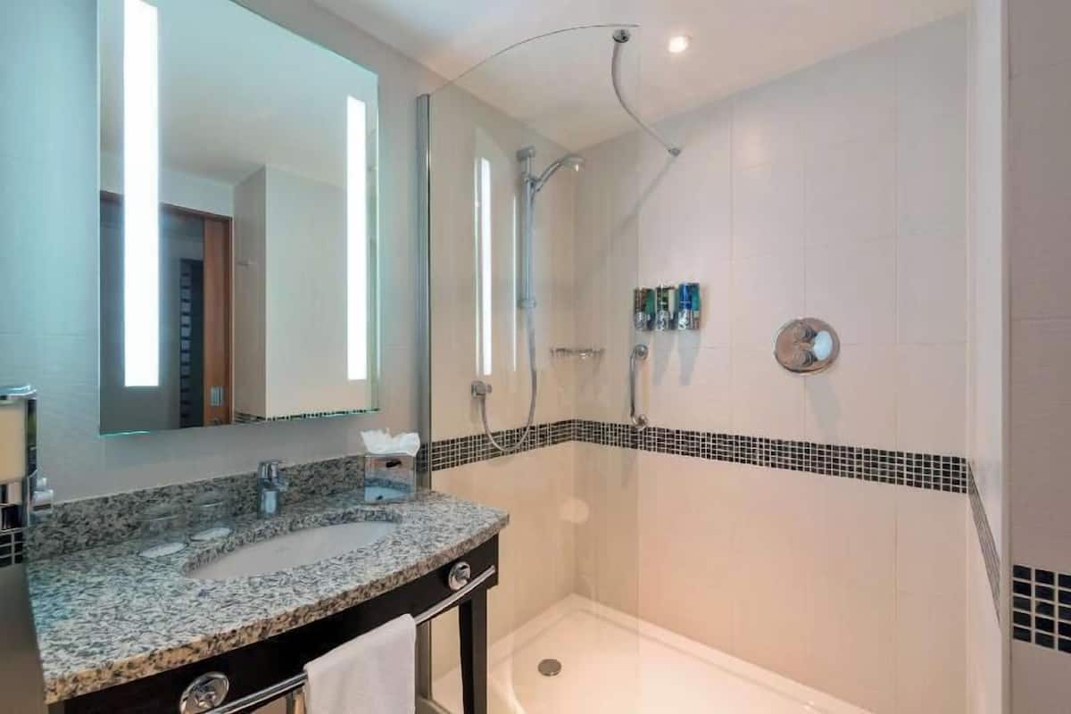 A modern bathroom with a granite countertop, a large mirror, a shower with a glass door, and a tiled wall with a black border.