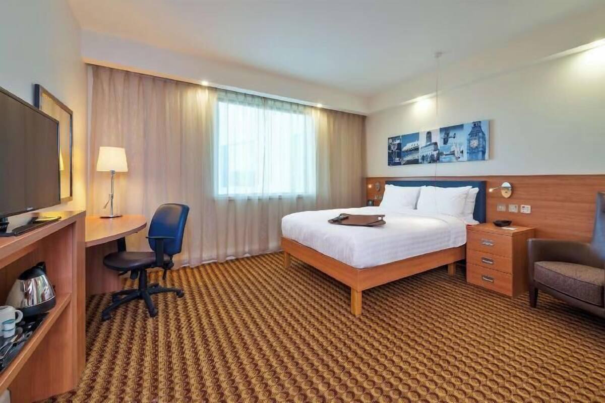 A hotel room with a bed, desk, chair, lamp, and a painting on the wall.