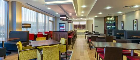 A modern hotel lobby with multiple seating areas, including tables and chairs, a reception desk, and a large window offering a city view.