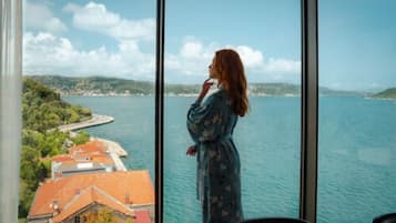 Una persona con vestido azul estampado de flores posa junto a un ventanal grande con vista a una masa de agua y un paisaje costero.
