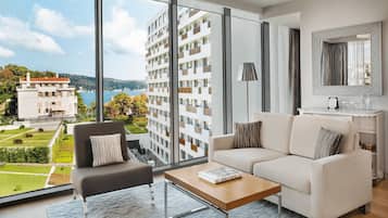Un moderno salón con sofá, sillón y mesa de centro, amplias ventanas que ofrecen vistas a un edificio y una masa de agua.