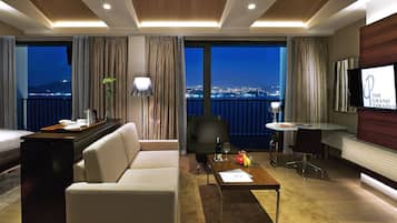 Habitación de hotel con sofá, una mesita con una botella y vasos, televisor y balcón con vista a la ciudad.
