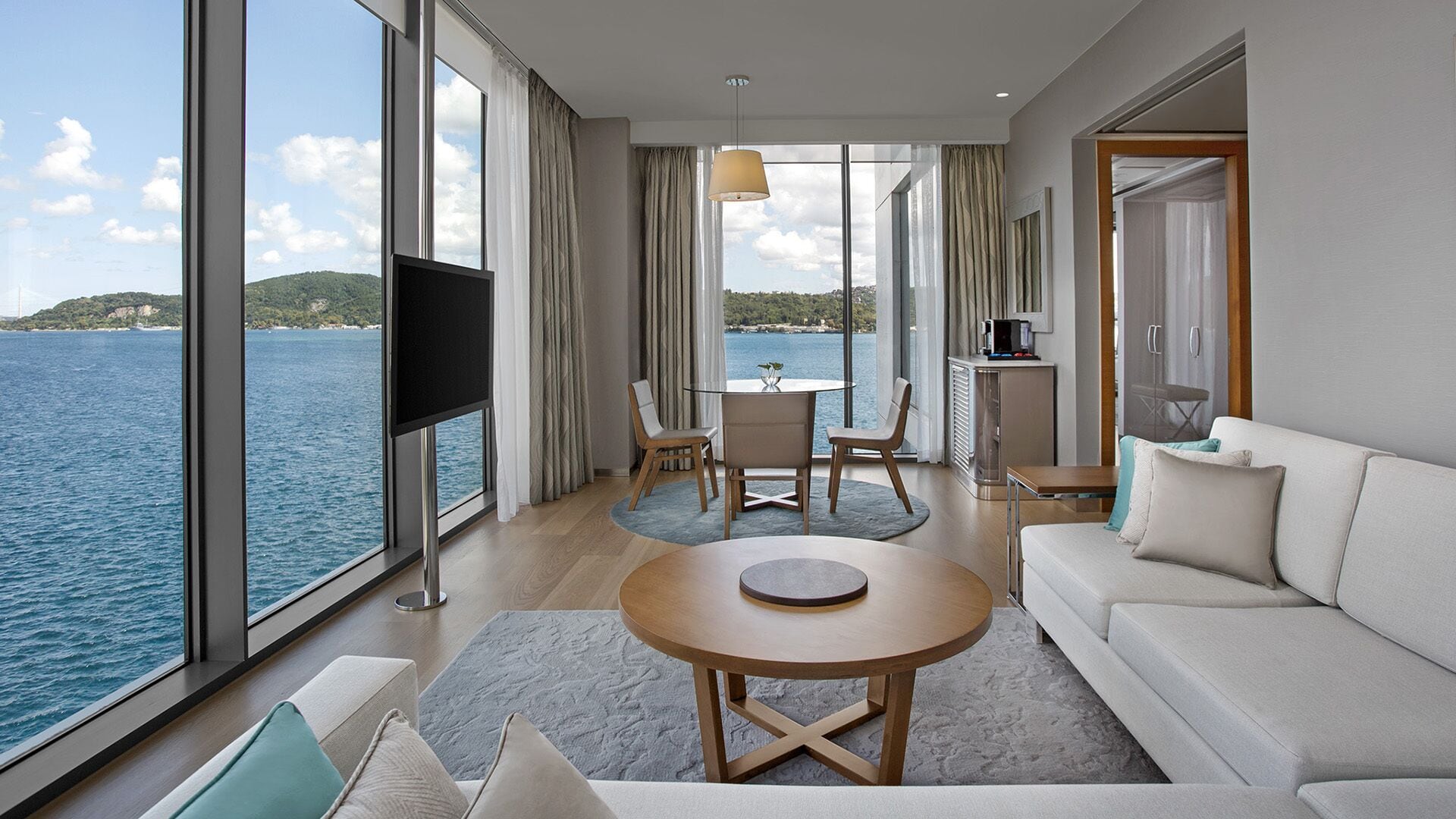 Habitación de hotel moderna con un amplio ventanal con vistas al mar, un televisor de pantalla plana, una mesa redonda de madera y un sofá.