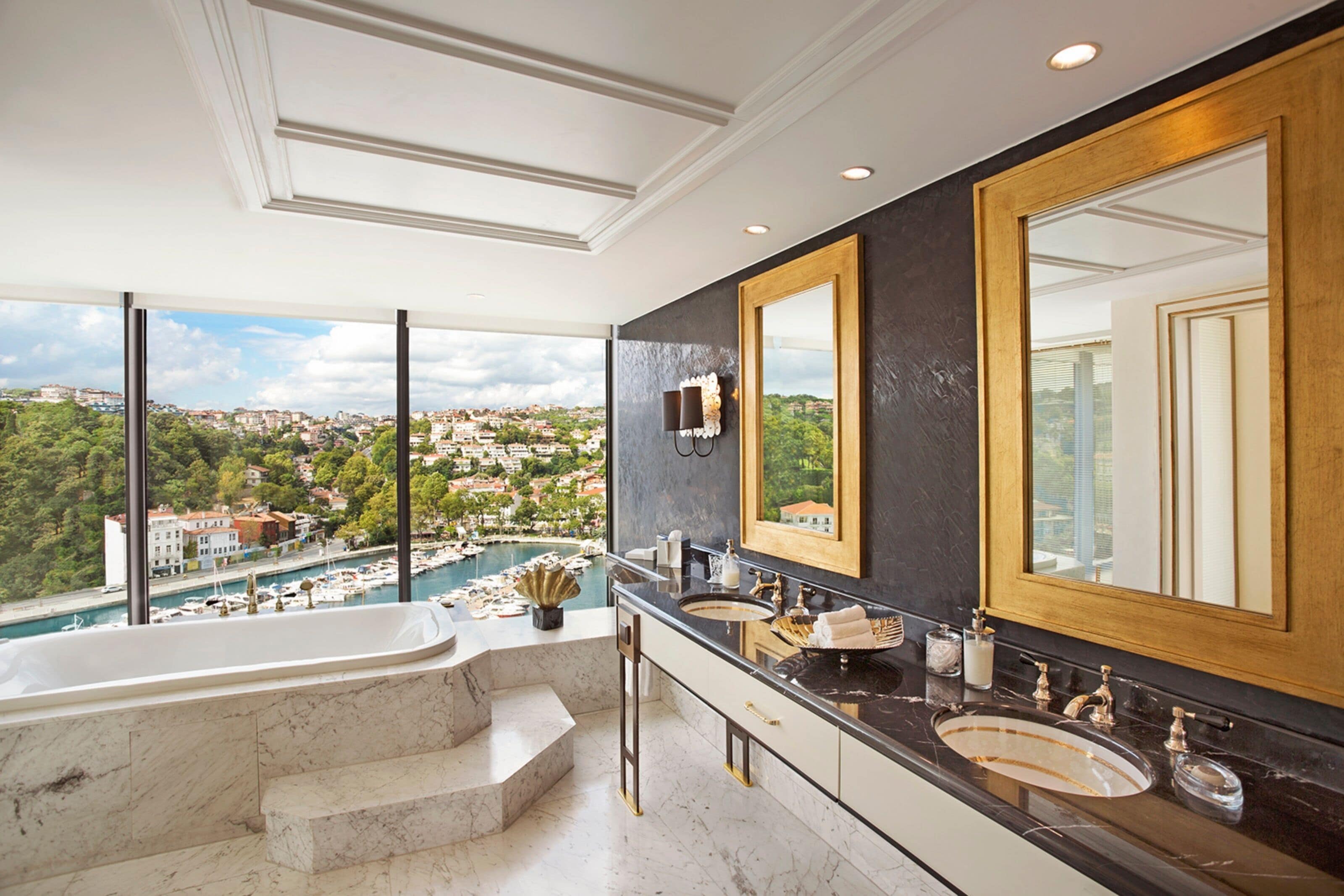 A luxurious bathroom with a large bathtub, two sinks, and a large mirror. It features a view of a marina and a cityscape.