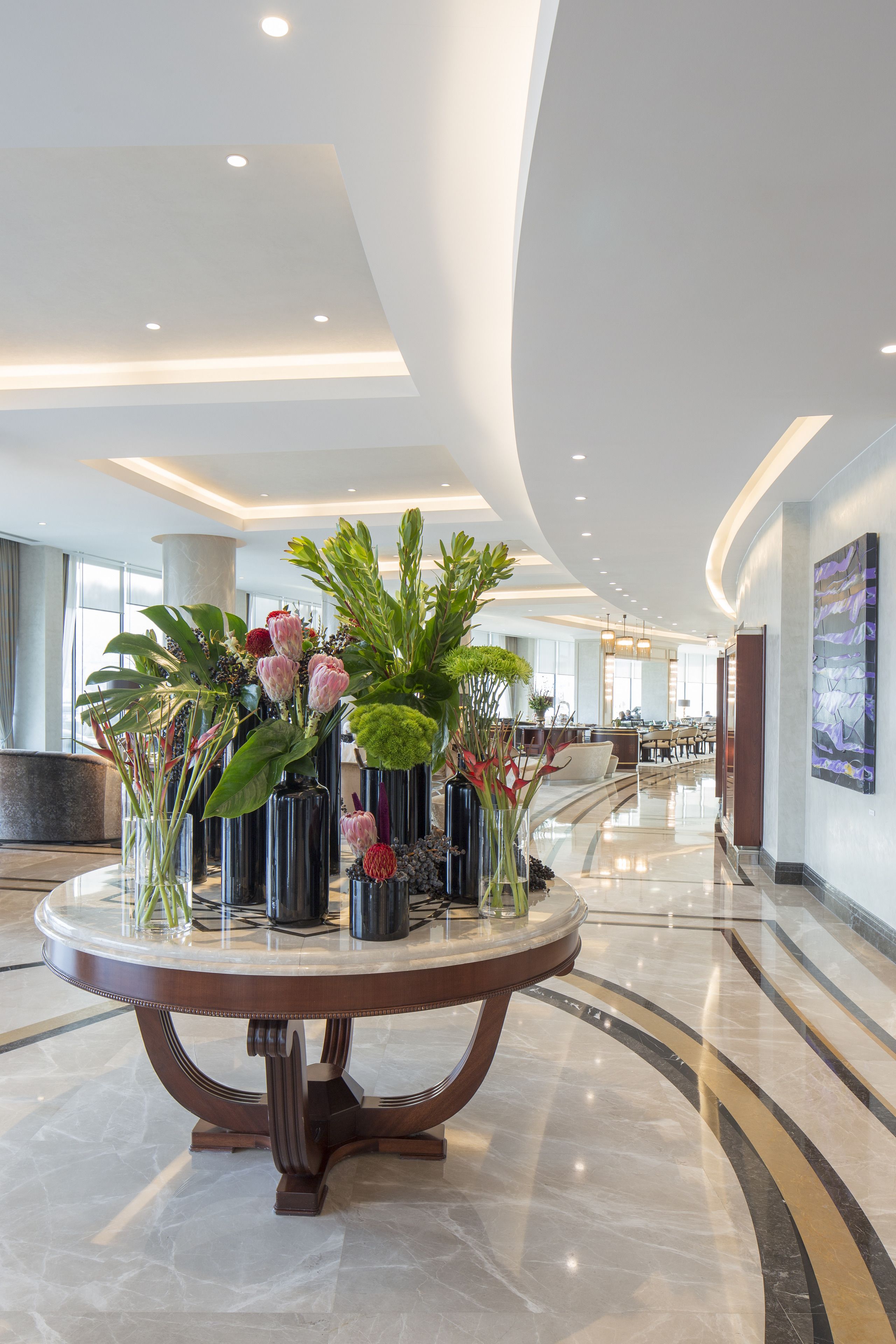 Un hall d’entrée moderne avec une table ronde en marbre entourée de vases de fleurs, un plafond incurvé doté d’un éclairage encastré et une vue sur la salle à manger en arrière-plan.