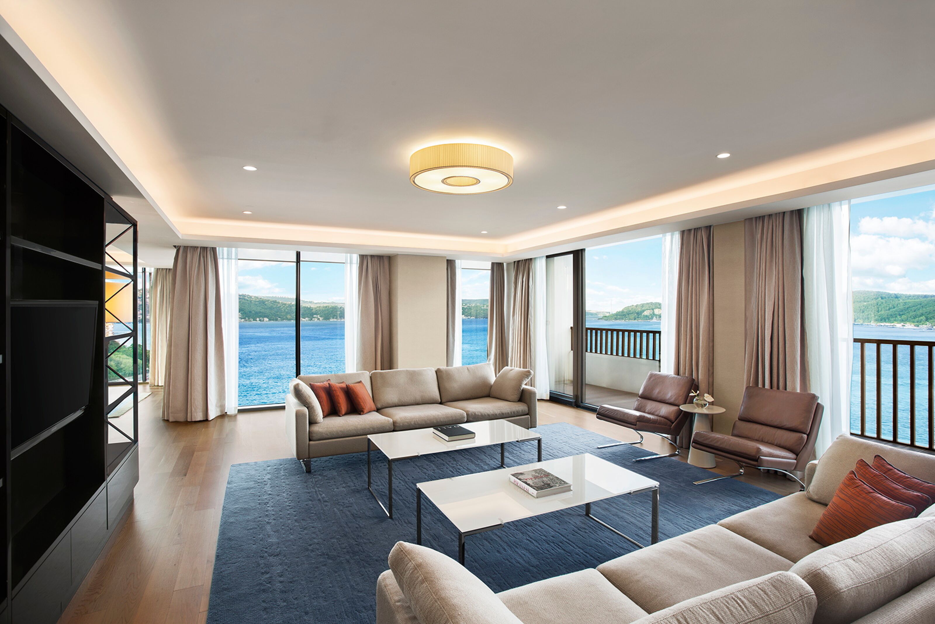 Ein geräumiges Wohnzimmer mit großem Fenster mit Blick auf ein Gewässer, einer Sofagarnitur, einem Couchtisch und einem Bücherregal.